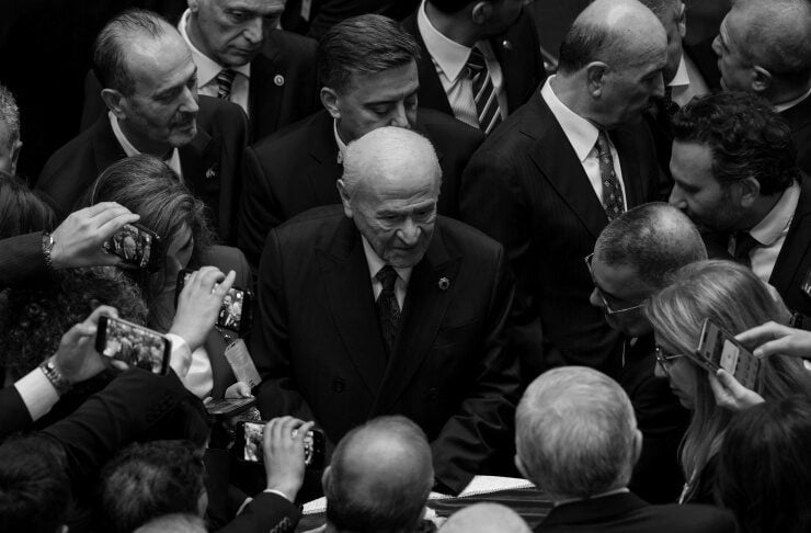 Elderly man in a dark suit surrounded by reporters and photographers at a crowded event in black and white.