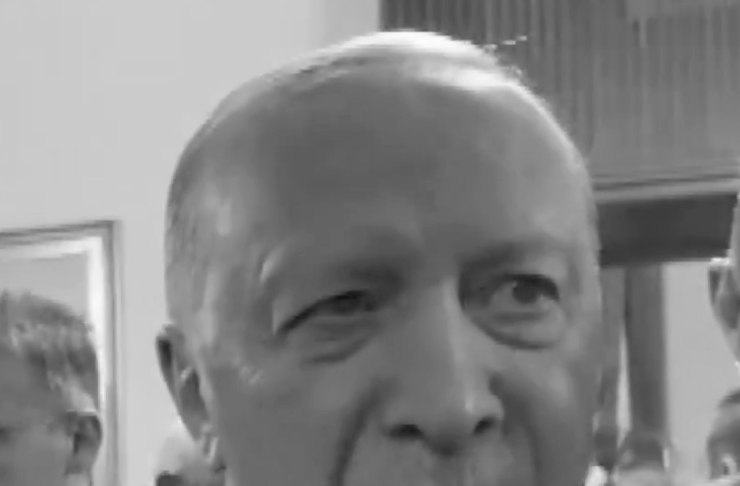 Grayscale close-up of an older man in a suit and tie speaking at a formal event, with a small flag lapel pin visible on his jacket.