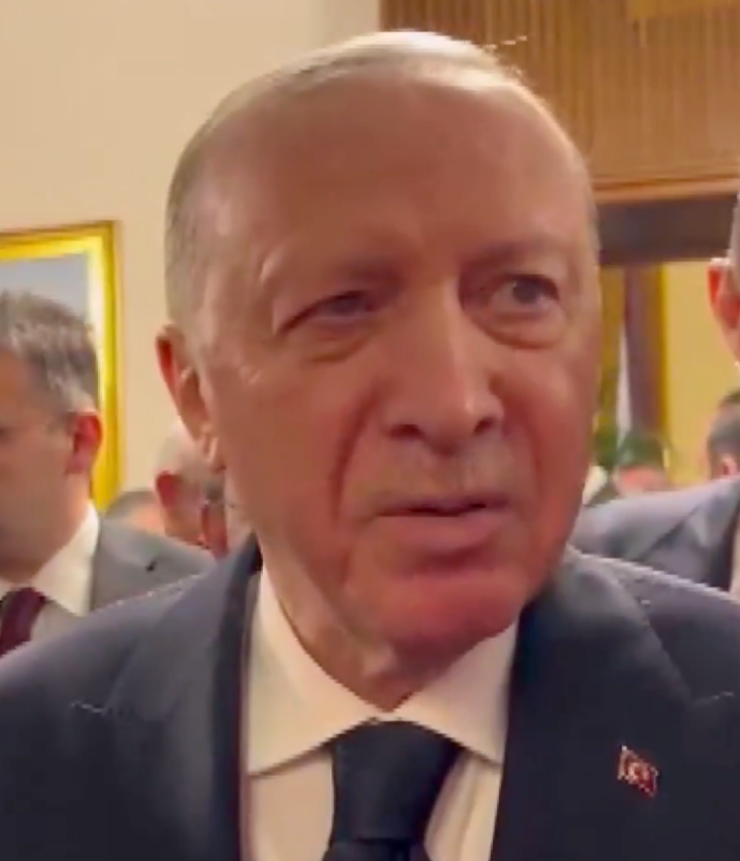Close-up of a gray-haired man in a dark suit and tie speaking at a formal indoor event, with a small flag pin on his lapel