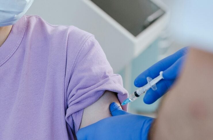 Close-up of a syringe injecting vaccine into a patient's upper arm as a healthcare worker wears blue gloves nearby, lavender shirt visible