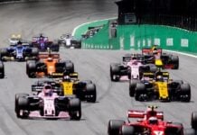 Several Formula 1 race cars speed along a circuit, with red, pink, and yellow cars leading. Green barriers line the track.
