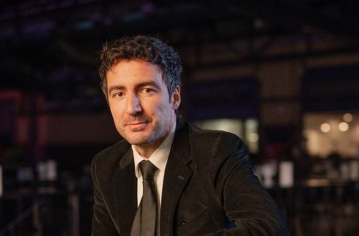 Man in a black suit and tie sits in a dimly lit venue, hands folded, looking at the camera.