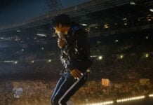 Concert performer on stage in a leather jacket, head down, performing to a crowded arena at night with bright lights overhead.