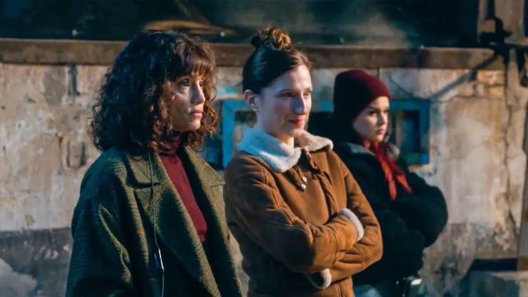 Three women stand side by side in an outdoor urban setting, wearing vintage coats and looking pensively ahead.