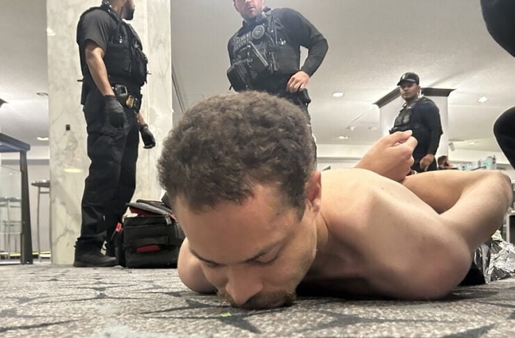 Man lying face down on carpet with arms restrained behind his back while armed security personnel stand nearby in a public indoor space.