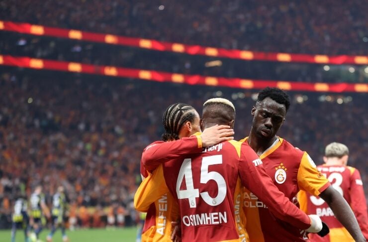 Group of Galatasaray players hug and celebrate on the pitch as fans cheer in a packed stadium in the background.