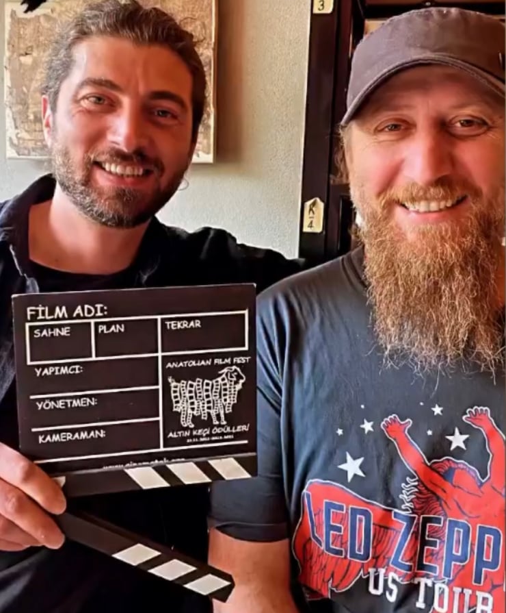 Two smiling men pose for a selfie; the man on the left holds a small film slate with blank fields and clapper marks visible at the bottom edge.