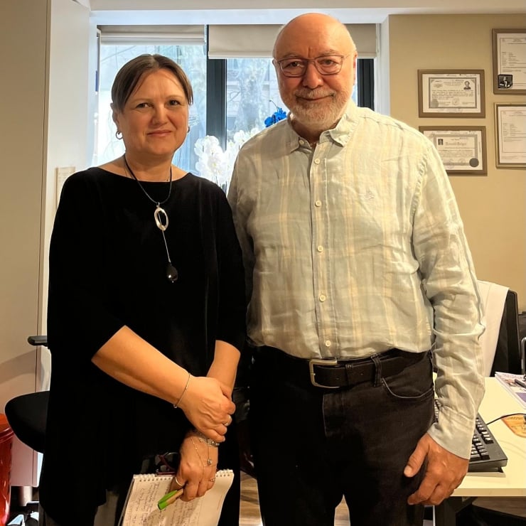 Two people stand side by side in an office, smiling at the camera with certificates on the wall behind them.