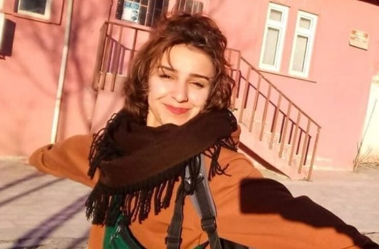 Young woman with short brown hair in an orange coat and dark scarf, arms outstretched, smiling outdoors near a pink building with stairs, wearing a backpack.