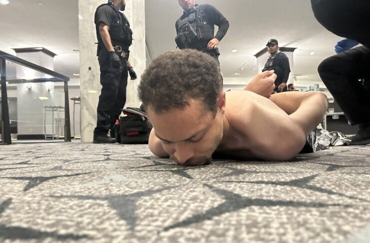 Shirtless man lying face down on a patterned carpet, surrounded by multiple armed officers in tactical gear in a bright indoor space.