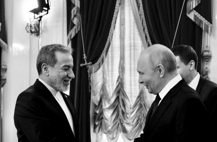 Two men in suits shake hands in a formal room with ornate curtains in the background.
