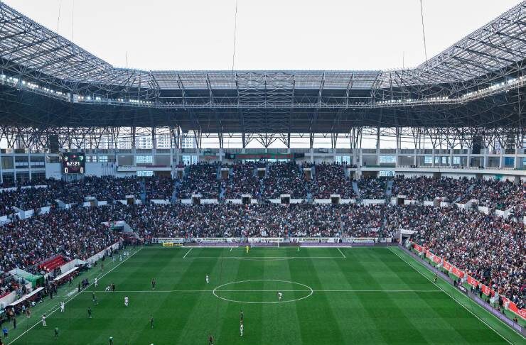 Aerial view of a packed soccer stadium with fans, green pitch, and players during a match near the goal area in the foreground.