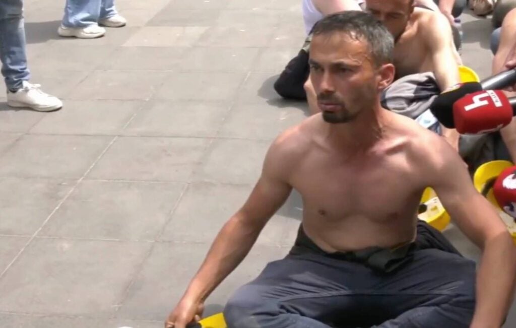 Shirtless man sits cross-legged on a paved surface as reporters with red microphones interview him nearby, with onlookers in the background.
