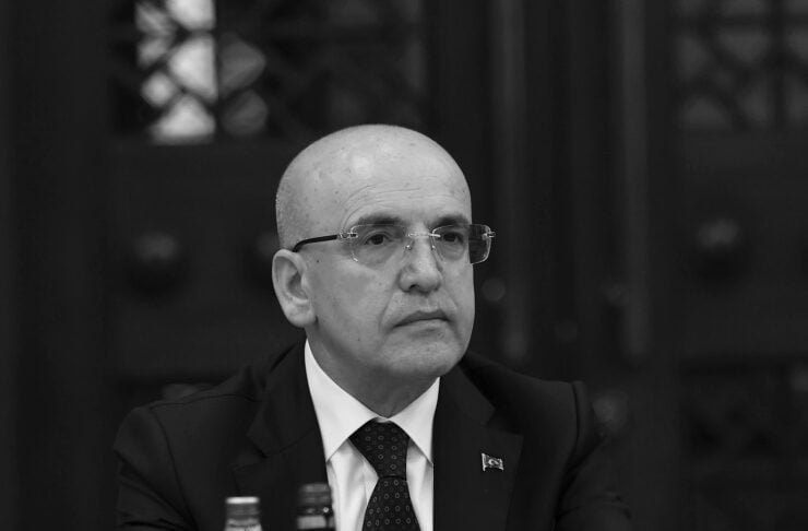 Bald man wearing glasses and a suit sits at a conference table with microphones and a nameplate in front, in a formal meeting setup.