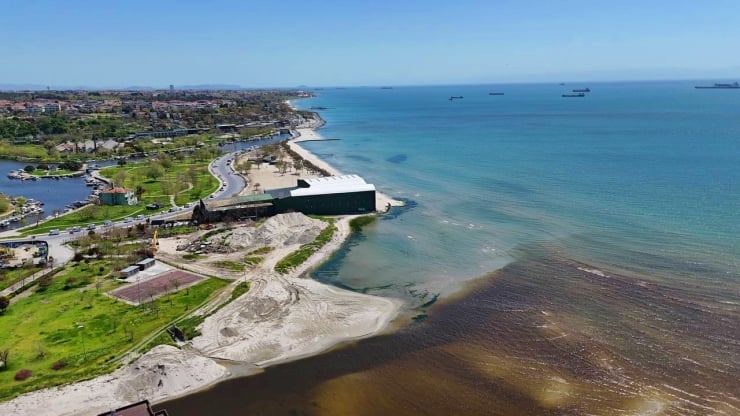 Aerial view of a coastal area with a sandy beach, green park, and a large dark building along the shore near calm blue water with ships on the horizon
