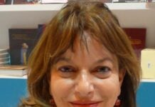 Close-up portrait of a smiling woman with light brown hair and red earrings, bookstore shelves in the background.