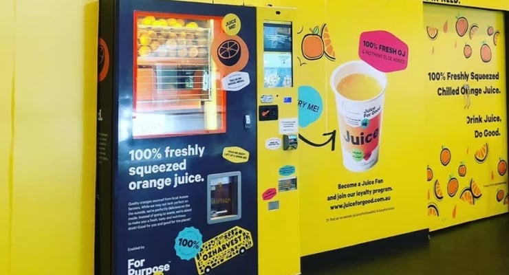 Bright yellow juice vending machine area promoting 100% freshly squeezed orange juice with orange graphics on the wall.