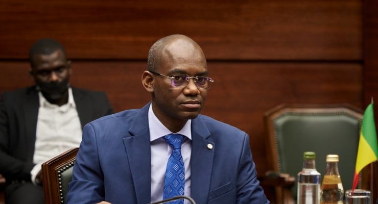 Man in a blue suit and patterned blue tie seated at a formal meeting table, with a flag in the background and water bottles nearby
