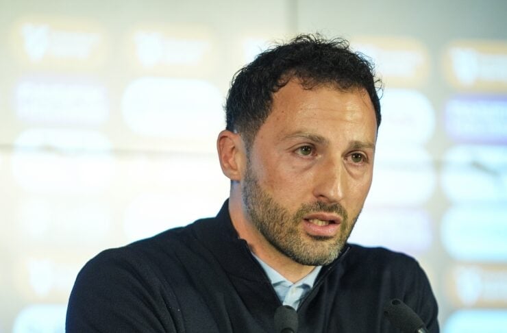 Man speaking at a press conference, seated behind microphones with a blurred backdrop of logos in the background.