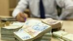 Businessperson in a shirt and tie counts large stacks of banknotes on a desk while writing notes nearby.