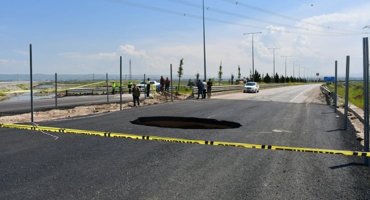 Road under construction with a large hole in the asphalt, yellow caution tape, construction workers, and a white car nearby; coastal landscape in the background