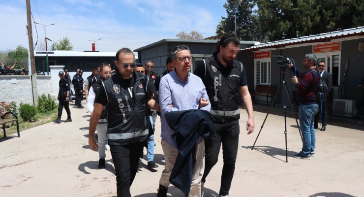 Man in a light blue shirt escorted by two officers in black vests; other officers and media filming in the background, outdoor facility with buildings.