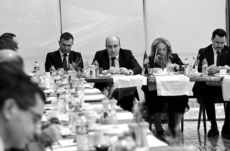 Formal meeting around a long table with officials; Turkish flag on the table and water bottles visible.