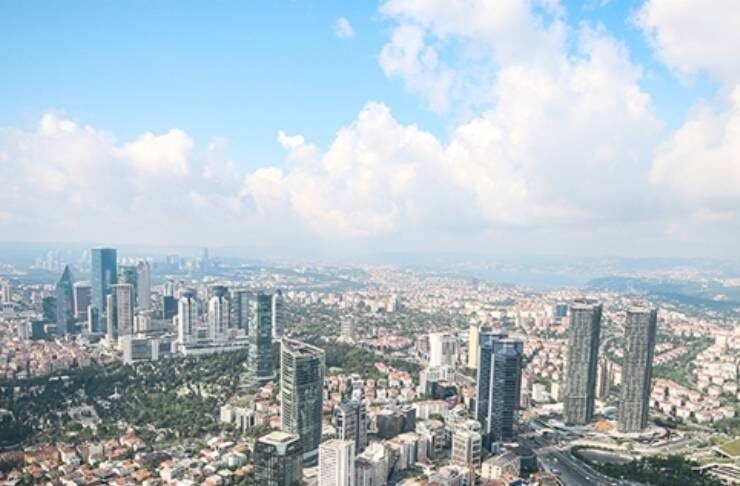 Aerial view of a modern city with tall skyscrapers, dense residential blocks, and a complex highway interchange under a blue sky.