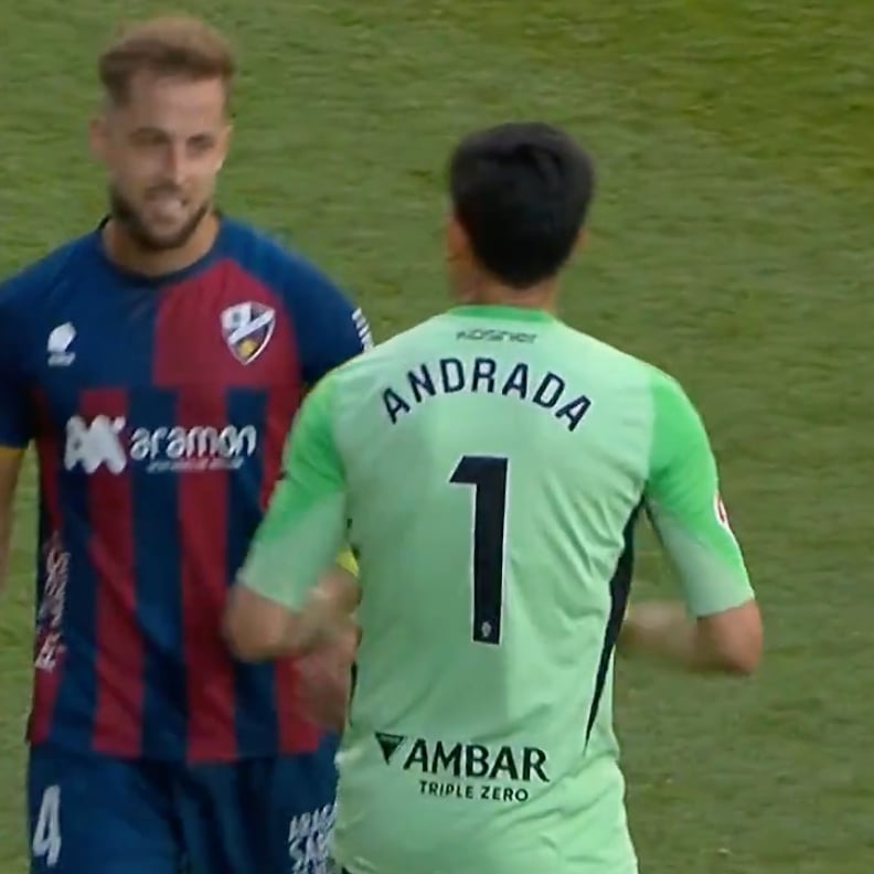 Two soccer players greet each other on a grass field: a pale green goalkeeper jersey with 'ANDRADA' and number 1 facing away, shaking hands with a blue-red kit opponent.