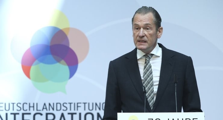 A middle-aged man in a dark suit speaks at a podium at a formal event, with a colorful circular logo in the background.