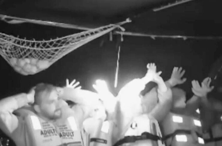 Group of people in life jackets on a small boat at night, raising hands in a panicked gesture.