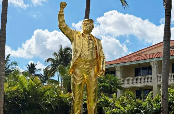 Gold-colored statue of a man in a suit with his right fist raised, atop a white pedestal in a tropical setting.