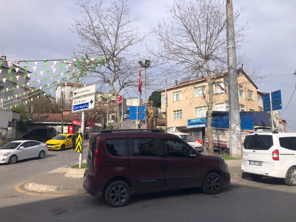 Busy urban intersection with a maroon van in the foreground, taxis, directional signs, leafless trees, and a bronze statue in the background.
