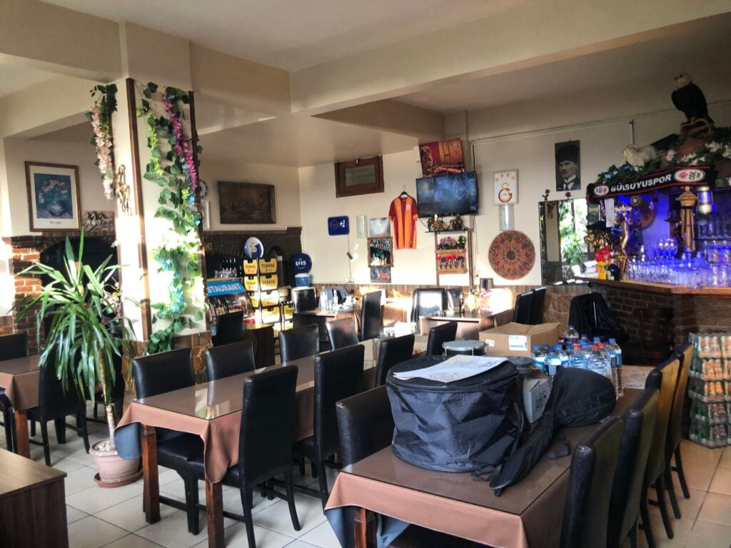 Cozy restaurant dining room with brown tablecloths, black chairs, and plants along a column; bar area with blue lighting on the right.