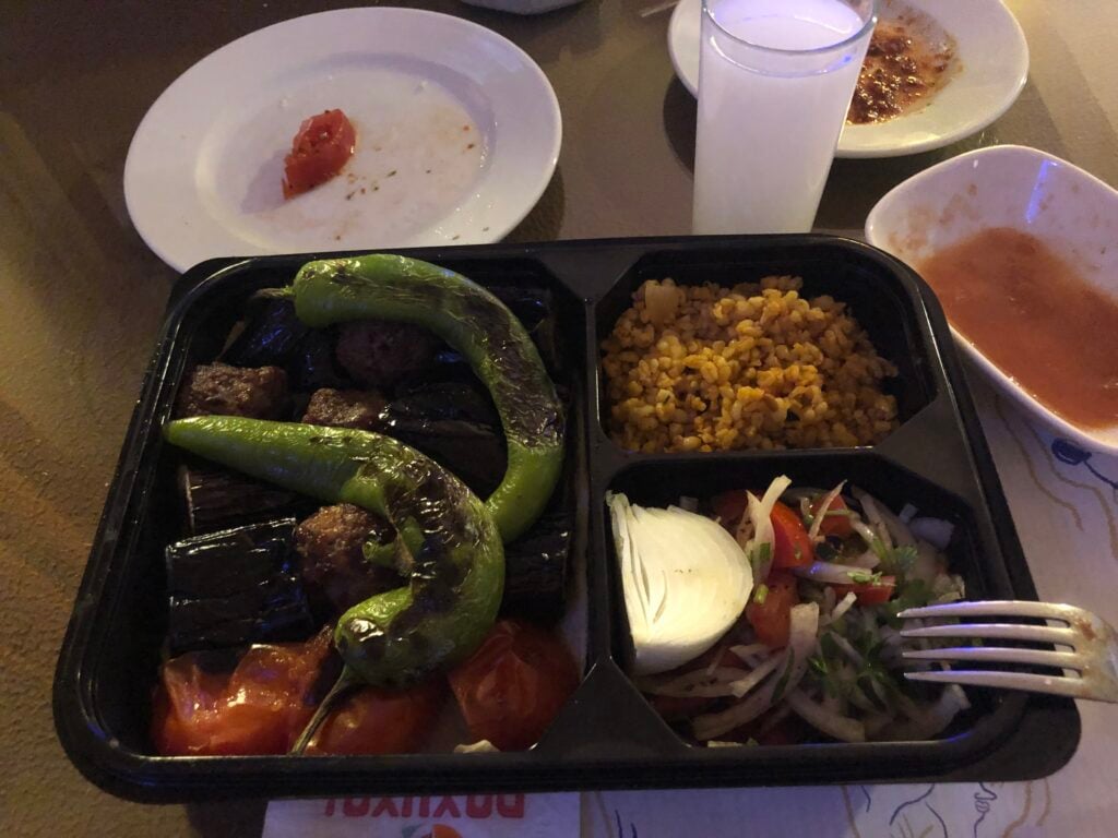 Bento box with grilled peppers, eggplant, tomatoes, and rice; side salad with onions and a lemon wedge, plus a sauce boat nearby.