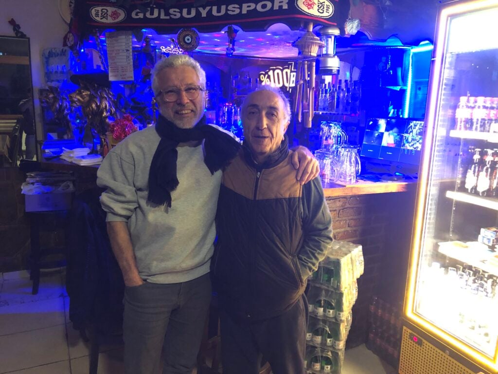 Two older men stand together in a bar, arms around each other, smiling under blue neon lighting near a drinks counter and fridge filled with bottles.