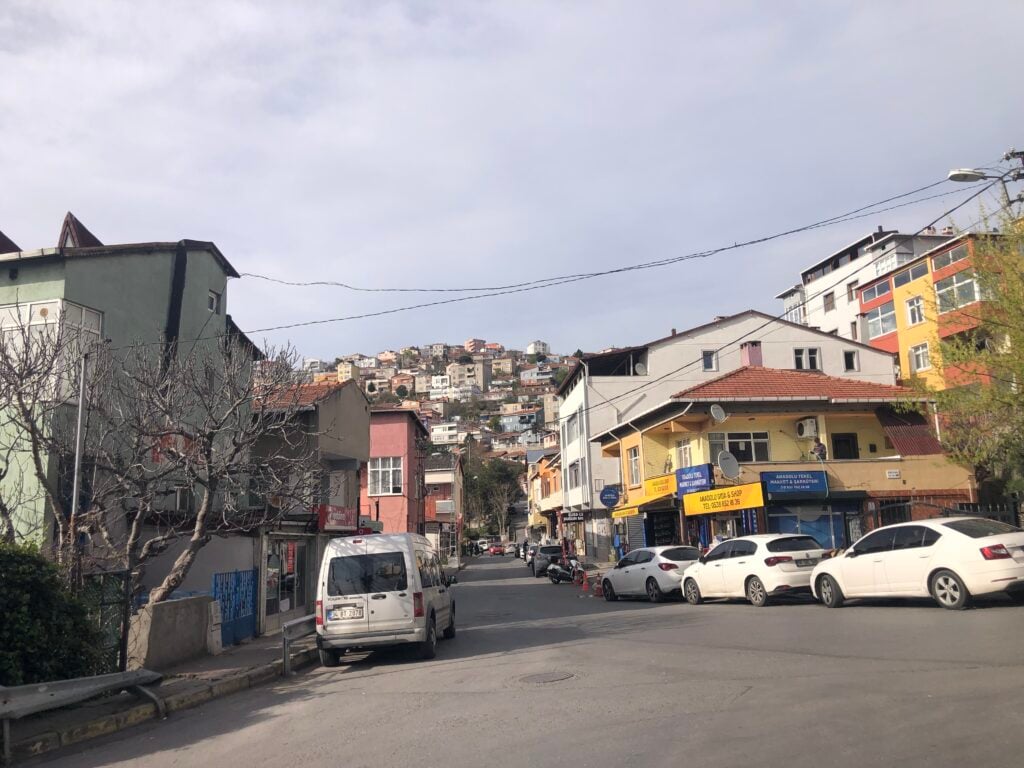 Sunny urban street in a hill town with colorful storefronts, parked cars, and a hillside neighborhood in the background.