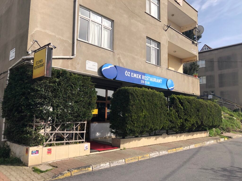 Facade of Öz Emek Restaurant with a blue sign over the entrance, beige apartment building, and trimmed hedges along the storefront.