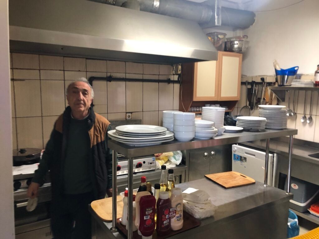 Older man in a brown-and-black jacket stands in a small commercial kitchen beside a stainless steel prep table with stacks of white plates.
