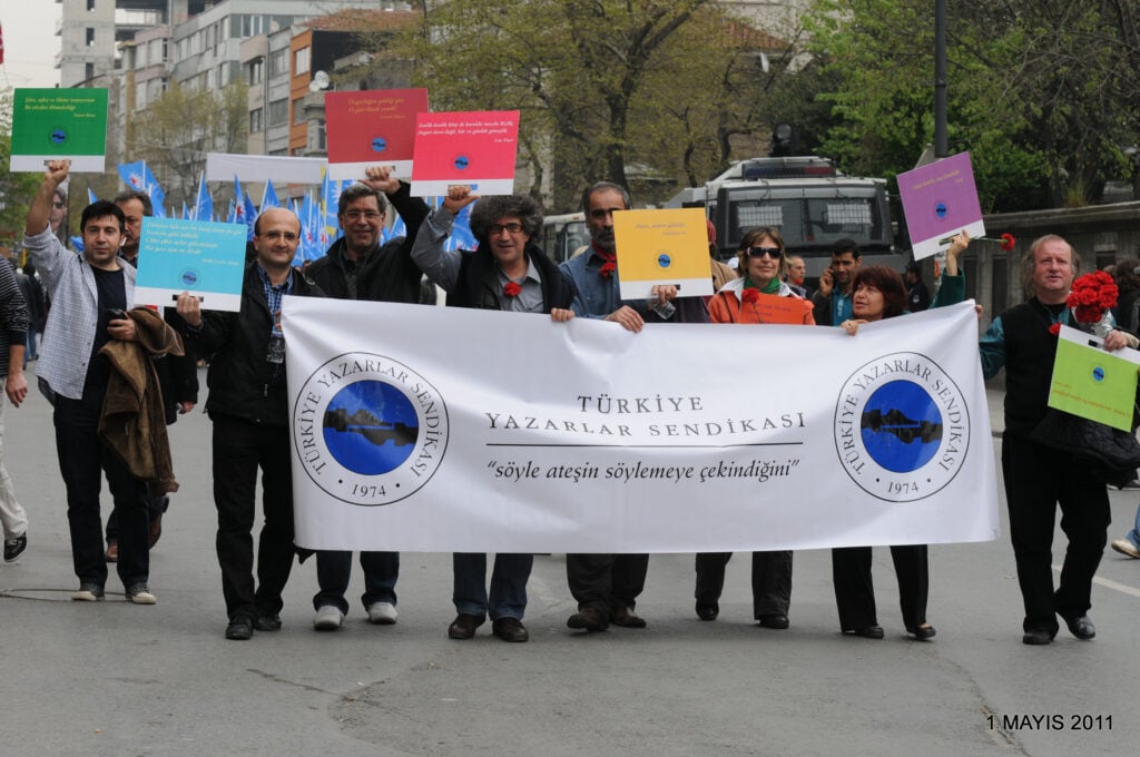 Group of protesters marching on a city street holding colorful placards and a large banner reading Türkiye Yazarlar Sendikası (Turkish Writers Union).