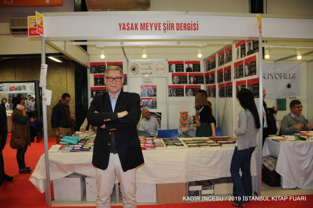 Man in blazer with arms crossed at a booth for 'YASAK MEYVE ŞİİR DERGİSİ' at the Istanbul Book Fair, with books on the table.