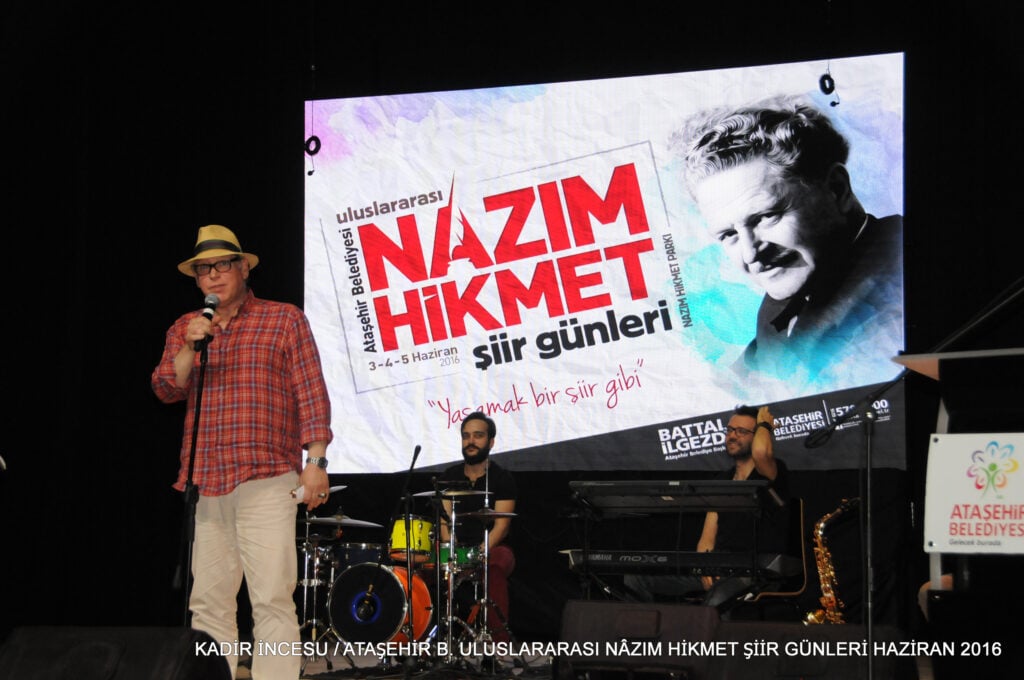 Man in a tan fedora and red plaid shirt speaks into a microphone on a stage, with a band and large poster behind him advertising Nazım Hikmet Poetry Days.