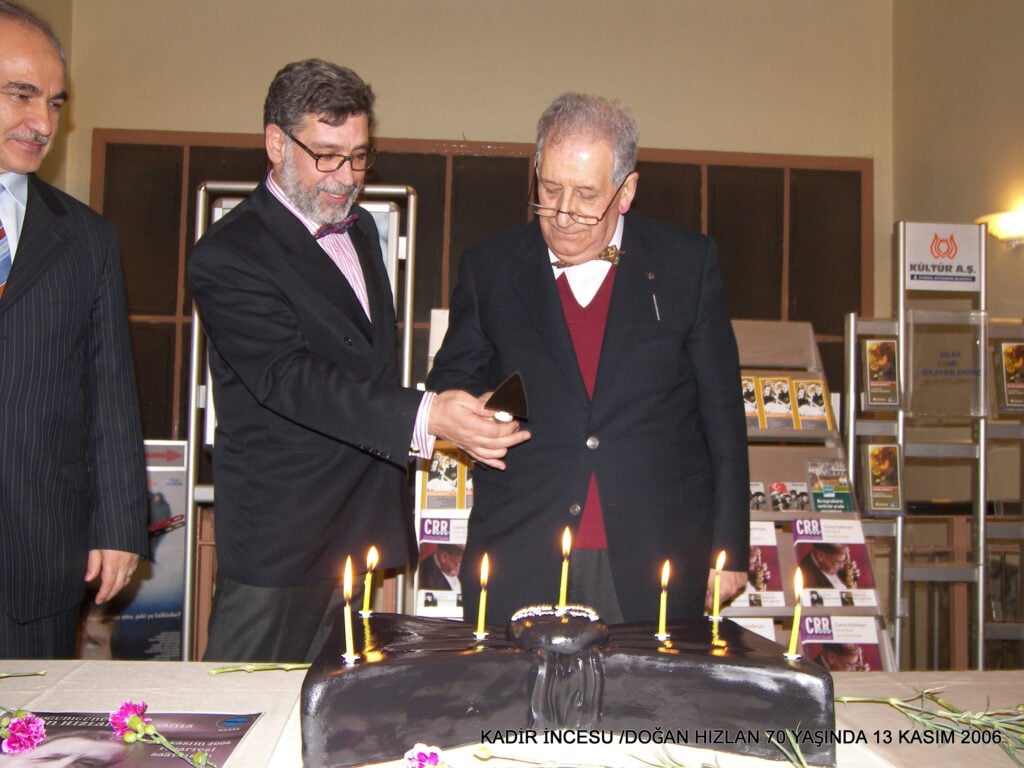 Two men in suits stand by a large chocolate cake with seven lit candles at a 70th birthday celebration for Doğan Hizlan (Nov 13, 2006). Versatile event photo text included at bottom edge reads the names and date.