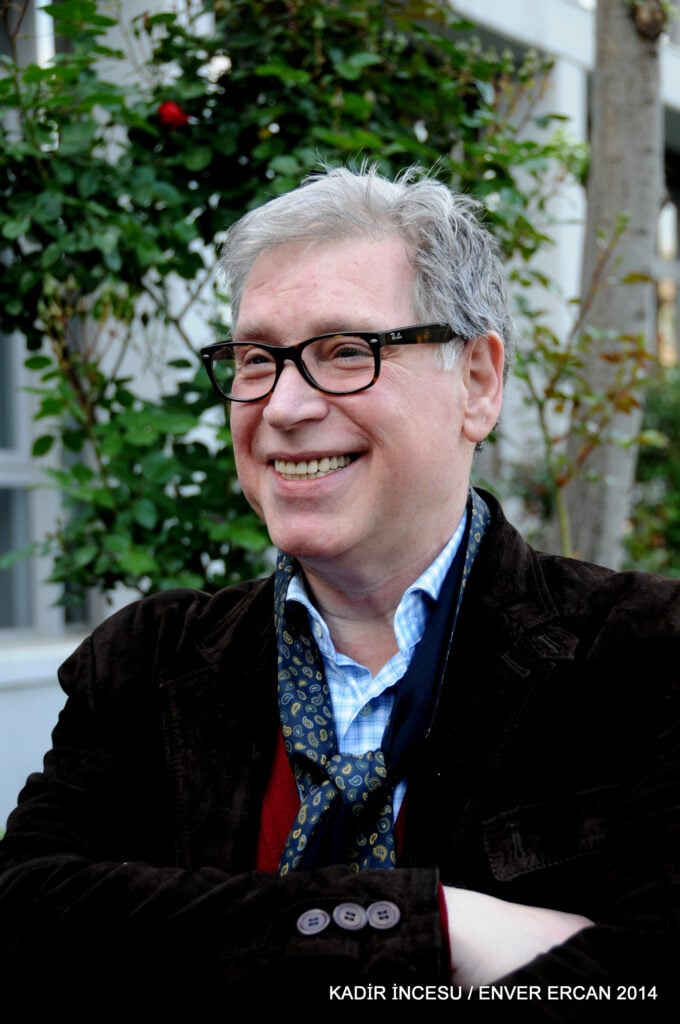 Smiling man with gray hair and black glasses outdoors, wearing a blue checkered shirt and dark blazer.