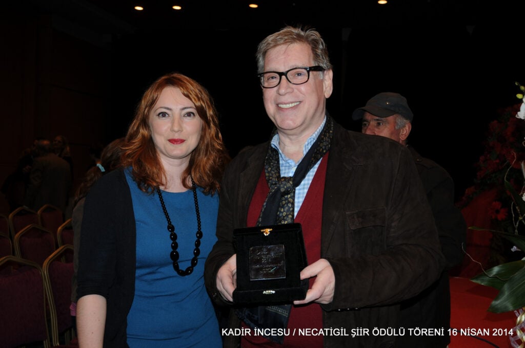 Two people pose for a photo at an awards event; a man in a brown jacket holds a small black velvet box while smiling beside a woman in a blue dress with a black beaded necklace.