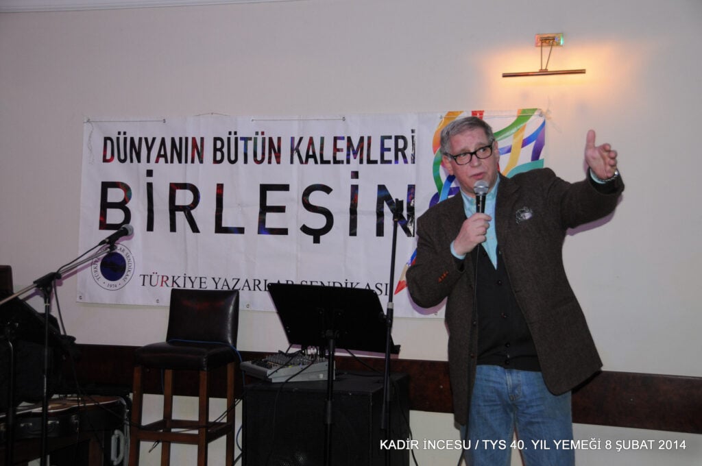 Speaker with microphone gesturing on stage in front of a banner, at a Turkish literary event.