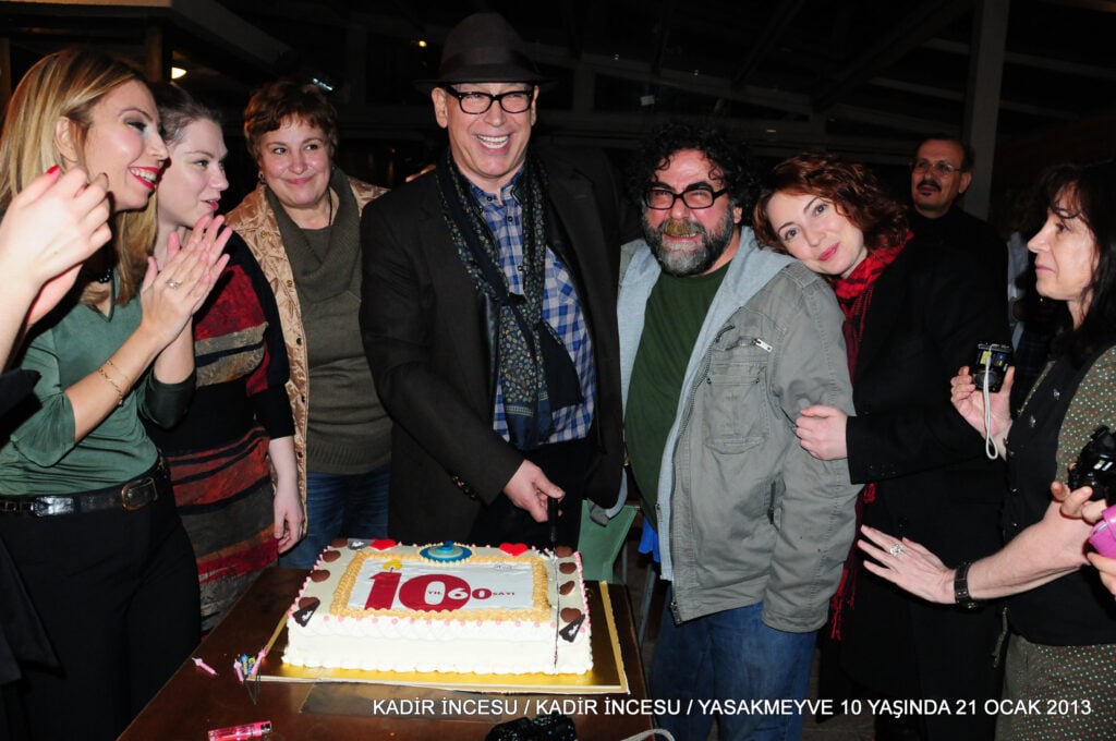 Group of adults gathered around a large decorated cake, smiling for a celebratory photo indoors.