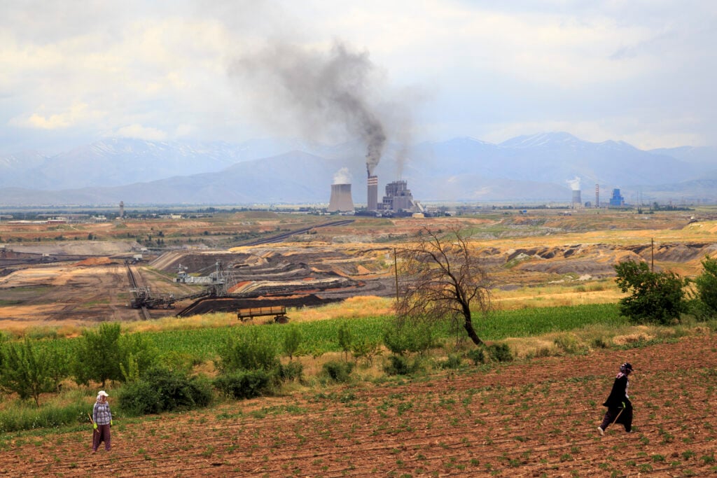 Industrial facility with tall smokestacks emitting dark smoke in a valley, mountains in the background; two people working in the foreground fields.