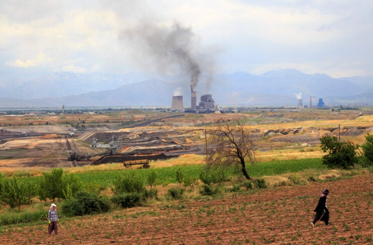 Industrial facility with tall smokestacks emitting dark smoke in a valley, mountains in the background; two people working in the foreground fields.