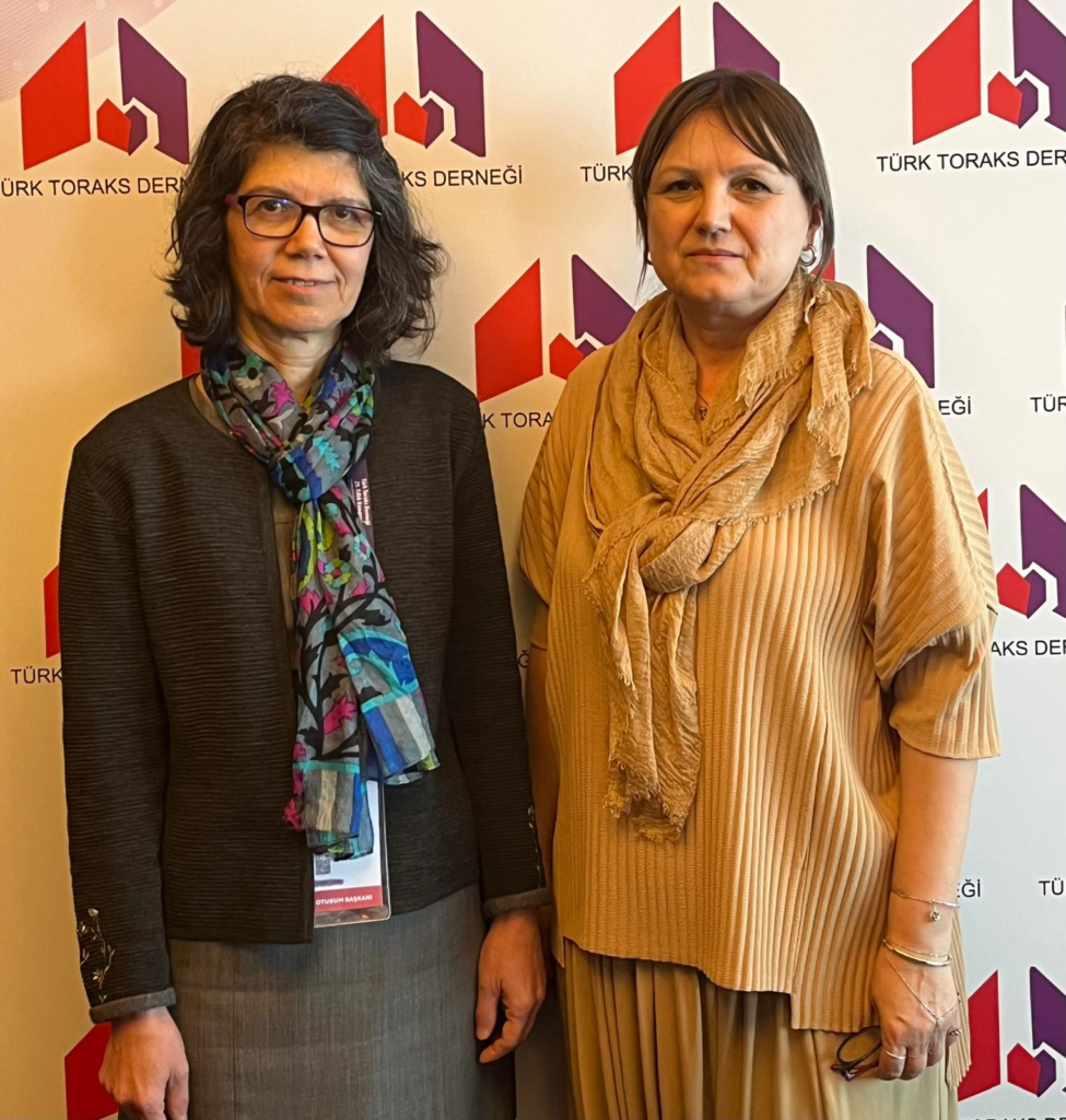 Two women standing side by side in front of a white backdrop with red and purple Turkish Thoracic Society logos; the left woman wears glasses, a dark jacket and a colorful scarf, and the right woman wears a peach pleated dress with a tan scarf.